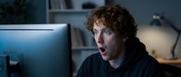 Shocked young man with curly reddish-brown hair and fair complexion stares at computer monitor in home office, expressing surprise or concern while working late at night.