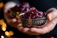 Hands holding ornate silver bowl filled with ruby red cranberry sauce against dark background with warm golden bokeh lights for holiday dining.