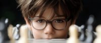 Young Asian boy wearing glasses peers intently over chess board during match, focused expression captures strategic thinking and concentration.