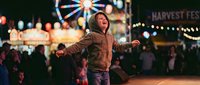 Young woman with arms outstretched enjoying nighttime carnival atmosphere with ferris wheel and colorful lights creating joyful celebration moment.