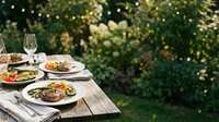 Outdoor dining table set with gourmet meals and wine glasses in garden setting with string lights and lush greenery for entertaining guests.