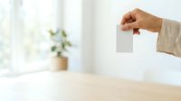 Hand holding blank white card or note in bright interior space with natural window light and potted plant, suitable for business messaging and promotional content.