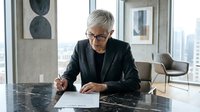 Senior professional woman with gray hair and glasses, wearing black blazer, signing documents at marble desk in modern high-rise office with city skyline view.
