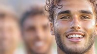 Young man with curly brown hair and beard, appearing to be of Mediterranean or Middle Eastern descent, smiling warmly at camera in professional portrait setting with blurred colleagues in background.