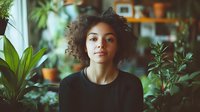 Young African American woman with natural curly hair in black top surrounded by green plants and natural light, creating serene indoor garden atmosphere.