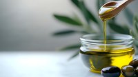 Golden olive oil pouring from wooden spoon into glass bowl with fresh olives and green leaves on white surface for cooking and culinary preparation.
