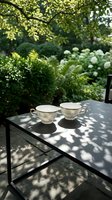 Two elegant white porcelain teacups on glass table in lush garden setting with dappled sunlight and green foliage creating peaceful outdoor dining atmosphere.