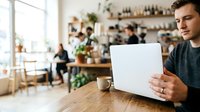 Young man of mixed ethnicity wearing casual dark clothing holds a blank tablet device while seated at wooden table in modern cafe with shelving, plants, and blurred customers in background.