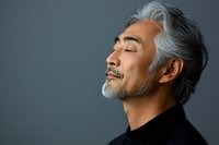 Serene mature Asian man with gray hair and beard in black turtleneck sweater against dark background, eyes closed with peaceful expression, side profile portrait.