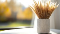 Dried wheat stalks in white textured ceramic vase against blurred autumn window background, creating minimalist home decor composition with natural materials.