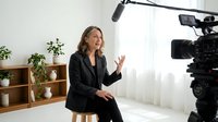 Middle-aged woman with long brown hair and light complexion, wearing black blazer, speaking during video production or interview recording in bright studio setting with professional camera equipment.