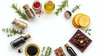 Gourmet food ingredients frame with herbs, spices, olive oil, chocolate, crackers and seasonings arranged in circle on white background for culinary design.