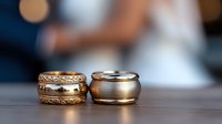 Close-up of elegant wedding rings on wooden surface with blurred bride and groom in background, symbolizing love and commitment at marriage ceremony.
