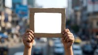 Hands holding blank white paper frame with brown border against blurred urban city street background for mockup design and advertising display purposes.