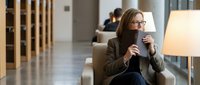 Professional businesswoman with glasses enjoying coffee break in modern office lobby, relaxing in comfortable chair with warm lighting.
