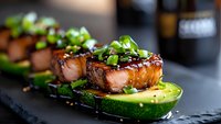 Grilled glazed salmon bites served on fresh avocado slices, garnished with chopped green onions and sesame seeds on black slate plate, modern food presentation.