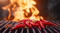 Fresh red chili peppers grilling on barbecue grate with dramatic flames and smoke in background, creating intense culinary atmosphere for spicy food preparation.