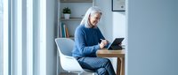 Senior Caucasian woman with gray hair using tablet computer while sitting in modern white chair at wooden desk near window in bright contemporary office.