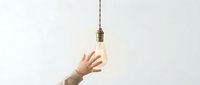 Hand reaching toward glowing vintage Edison bulb hanging from rope cord against white background symbolizing innovation and creative ideas.