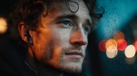 Young Caucasian man with curly hair looking thoughtfully through rain-covered window with colorful city lights bokeh background at night.