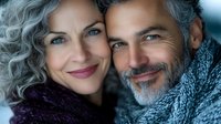 Mature couple in winter clothing sharing intimate moment outdoors, with gray and silver hair, warm smiles, and cozy knit scarves. Close-up portrait showing genuine connection.