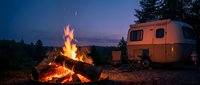 Cozy campfire glowing at dusk with vintage travel trailer in peaceful wilderness setting. Perfect for outdoor adventure and camping lifestyle content.