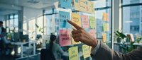 Professional pointing at colorful sticky notes on glass wall during brainstorming session in modern office environment with colleagues collaborating in background.