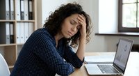 Exhausted businesswoman with curly hair experiencing work stress and fatigue while working on laptop in modern office environment.