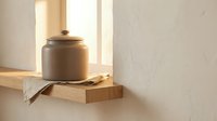 Minimalist ceramic jar with wooden lid on natural wood shelf against textured wall in warm sunlight for home decor and kitchen storage solutions.