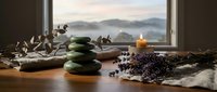 Spa wellness setup with stacked green stones, burning candle, dried lavender and eucalyptus branches on wooden surface near window with mountain view.
