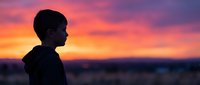 Young boy silhouette against vibrant sunset sky with pink and orange clouds, peaceful evening moment outdoors for lifestyle and family content.