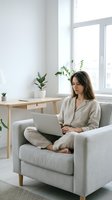 Young woman working from home on laptop computer sitting in comfortable armchair by window in bright modern living room with plants for remote work lifestyle.