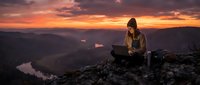 Woman with long dark hair and light skin tone, wearing casual outdoor clothing, sitting on a mountain peak with laptop during golden hour sunset, working remotely with scenic valley and river below.