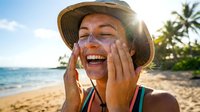 Young woman applying sunscreen on tropical beach vacation with palm trees and ocean background, sun protection skincare routine for summer travel.