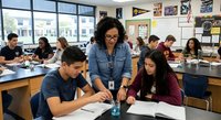 African American female teacher helping diverse high school students with assignments in modern classroom with large windows and educational posters on walls.