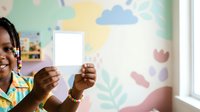 Young African American female child holding blank white card in playful classroom setting with colorful cloud and nature wall decorations, suitable for educational materials and children's content.