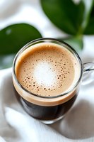 Fresh espresso with creamy foam in glass cup against blurred green leaves background, close-up view showing rich crema and tiny bubbles on surface.