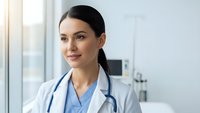 Professional female doctor in white coat with stethoscope smiling confidently in modern medical office setting for healthcare and medicine concepts.