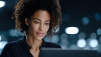 Professional African American businesswoman with curly hair working on laptop in modern office environment with city lights background at night.