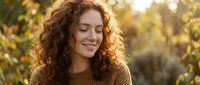 Young woman with curly red hair smiling peacefully in autumn sunlight wearing cozy sweater for lifestyle and wellness content.