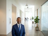 Male professional in dark business suit and blue tie standing in modern office corridor with glass partitions, potted plant, and blurred colleagues in background. Corporate workplace environment.