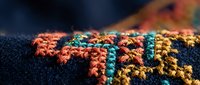Colorful embroidered textile with intricate thread work featuring orange, teal and yellow patterns on dark fabric for fashion and craft projects.