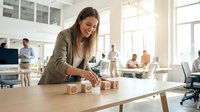 Young woman with long brown hair and light skin tone, wearing business casual attire, arranging wooden blocks with icons in modern office environment with colleagues working in background.