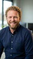 Smiling middle-aged Caucasian businessman with curly blonde hair and beard in navy shirt at modern office workspace for corporate headshot photography.