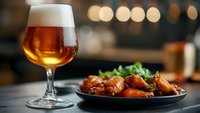 Glass of craft beer with frothy head alongside spicy chicken wings garnished with fresh herbs on dark plate, bokeh lights background creates cozy pub atmosphere.