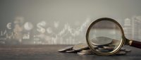 Financial analysis concept with magnifying glass examining coins against blurred stock market charts and data visualization background for investment research.