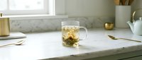 Glass mug with herbal tea and floating herbs on marble countertop in bright kitchen workspace, suitable for beverage, wellness, and lifestyle design projects.