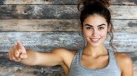 Young Hispanic woman in gray sports tank top showing thumbs up gesture with bright smile against rustic wooden wall background, wellness and fitness concept.