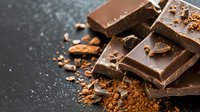 Dark chocolate pieces with cocoa powder scattered on black background, close up view showing texture and layers of broken chocolate bars for food photography.