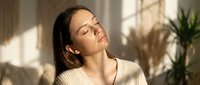 Young woman with closed eyes enjoying warm sunlight streaming through window in cozy home interior, expressing peace and relaxation in comfortable sweater.
